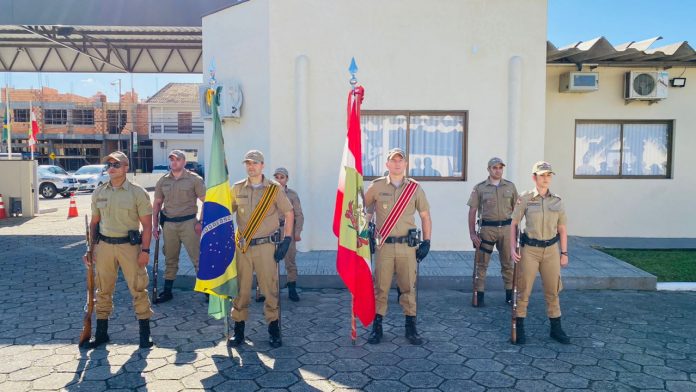 #PraCegoVer Na foto, oito policiais militares estão em formação. Dois deles, no centro, seguram bandeiras do Brasil e de Santa Catarina