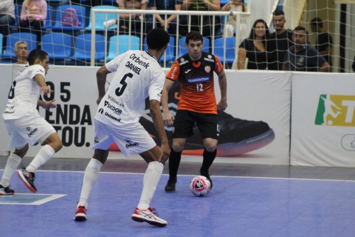 #PraCegoVer Na foto, dois jogadores de futsal com uniforme branco e um atleta com traje laranja e preto disputam a posse de bola