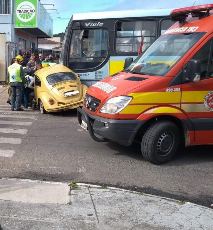 #PraCegoVer Na foto, bombeiros militares assistem uma mulher que ficou machucada após uma colisão entre o seu carro, um fusca amarelo, e um ônibus escolar. Em primeiro plano está a ambulância do Corpo de Bombeiros