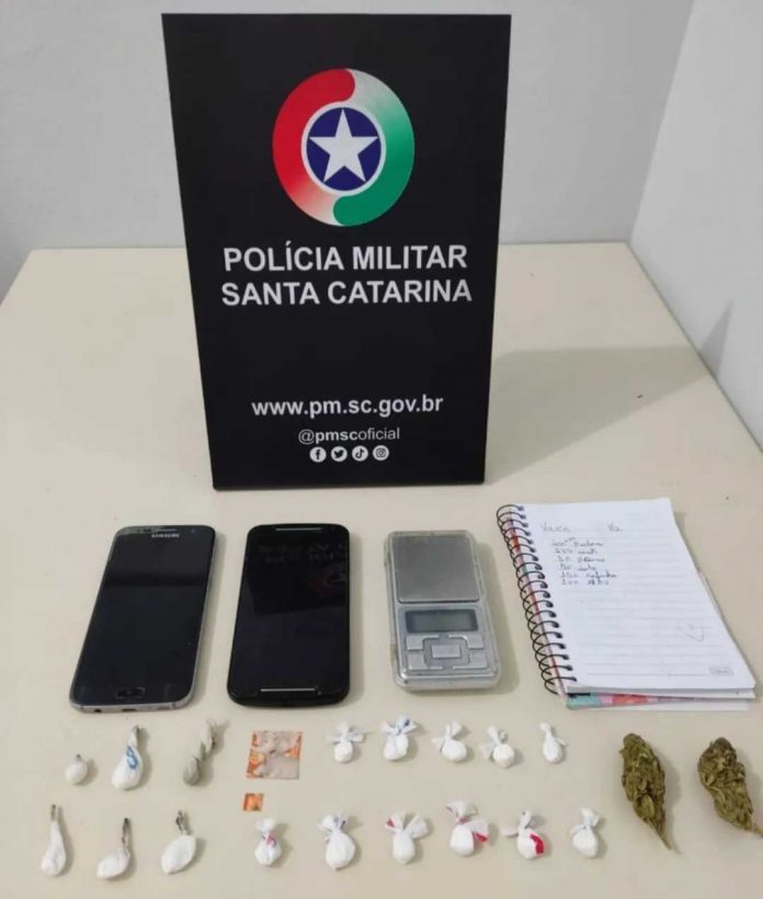 #ParaTodosVerem Na foto, em cima de uma mesa há uma placa com o símbolo da Polícia Militar, dois aparelhos de telefone celular pretos, uma balança de precisão, um caderno com anotações e porções de LSD, MDMA, skunk e cocaína