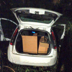 #ParaTodosVerem Na foto, um carro branco, abandonado em uma área de mata. No porta-malas duas caixas cheias com produtos roubados