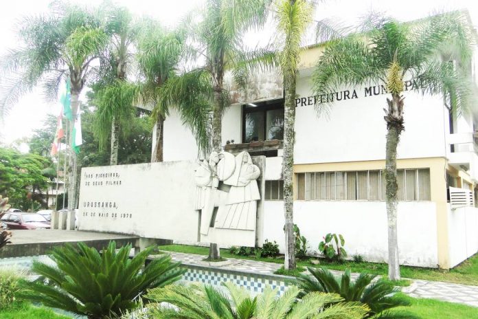 #PraCegoVer Na foto, um prédio branco, com um painel de cimento e plantas verdes na frente. No local funciona a sede da Prefeitura de Urussanga