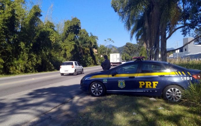 #PraCegoVer Na foto, um policial rodoviário federal fiscaliza uma rodovia. Ele está ao lado de uma viatura, às margens da estrada