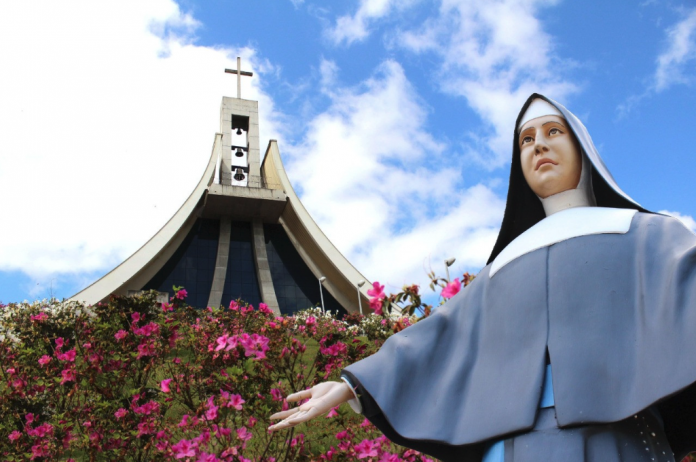 #ParaTodosVerem Na foto, uma imagem da santa Paulina, atrás há flores e uma igreja