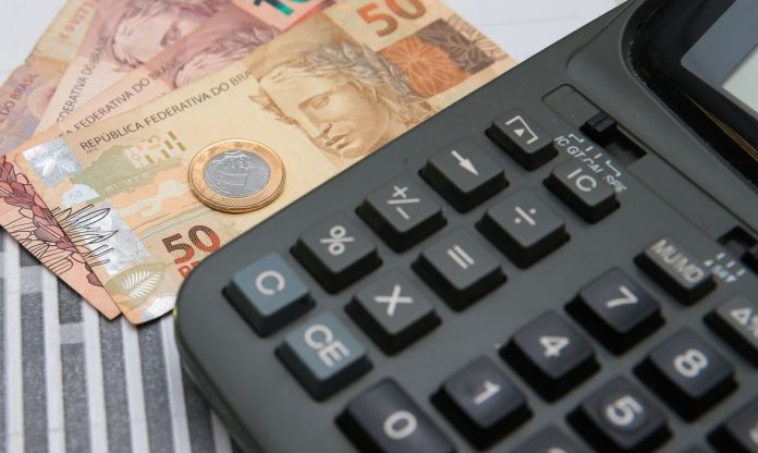 #ParaTodosVerem Na foto, em cima de uma mesa há uma calculadora, algumas cédulas de dinheiro e uma moeda