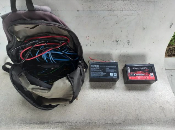 #ParaTodosVerem Na foto, em cima de um banco há uma mochila preta com vários fios elétricos e, ao lado, duas baterias