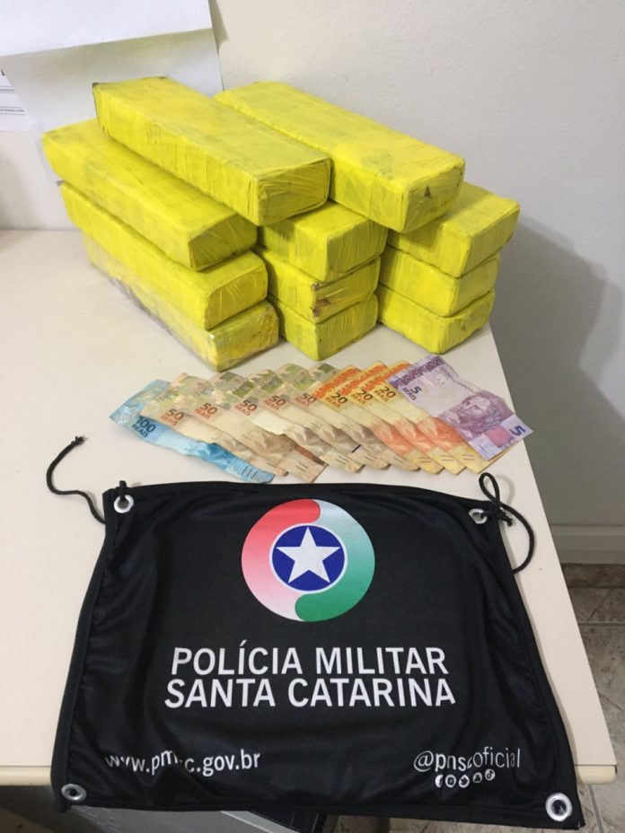 #ParaTodosVerem Na foto, em cima de uma mesa há 11 tabletes amarelos de maconha, cédulas de dinheiro e uma bandeira com o símbolo da Polícia Militar