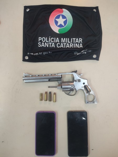 #ParaTodosVerem Na foto, uma bandeira com o símbolo da Polícia Militar, um revólver prata, quatro munições e dois aparelhos de telefone celular