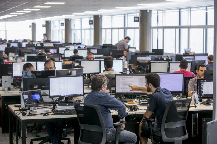 #ParaTodosVerem Na foto, uma sala grande, cheia de computadores e pessoas trabalhando