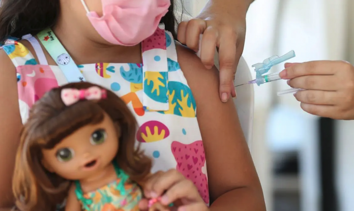 #ParaTodosVerem Na foto, uma menina é vacinada. Ela usa máscara cor de rosa e segura uma boneca
