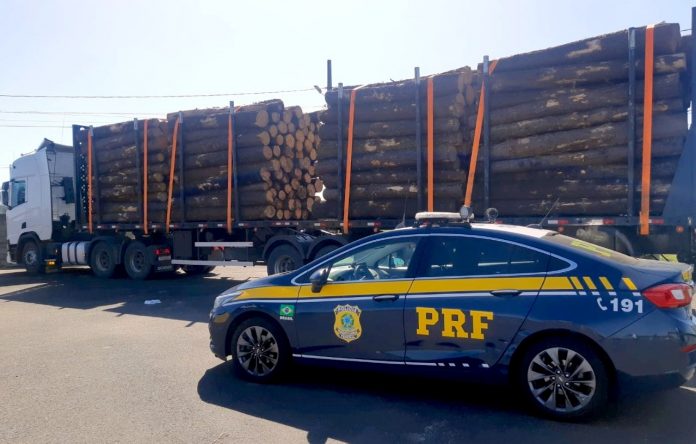 #ParaTodosVerem Na foto, uma viatura da Polícia Rodoviária Federal. Atrás há uma carreta com uma carga de toras de madeira