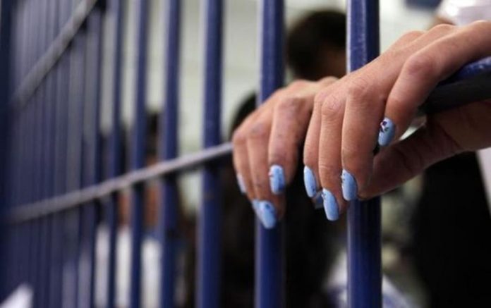 #ParaTodosVerem Na foto, uma mulher com as mãos apoiadas na grade de uma prisão