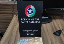 #ParaTodosVerem Na foto, em cima de uma mesa há uma placa com o símbolo da Polícia Militar, porções de drogas, um telefone celular preto e algumas cédulas de dinheiro