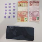 #ParaTodosVerem Na foto, comprimidos de ecstasy, um aparelho de telefone celular preto com a tela danificada e alguns cédulas de dinheiro