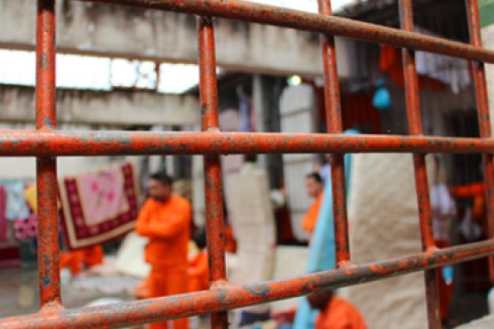 Imagem mostra presos, com uniformes laranja, fora de foco. Grades em primeiro plano.