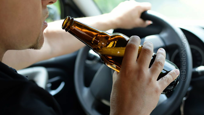 Foto mostra condutor ao volante segurando garrafa longneck de cerveja